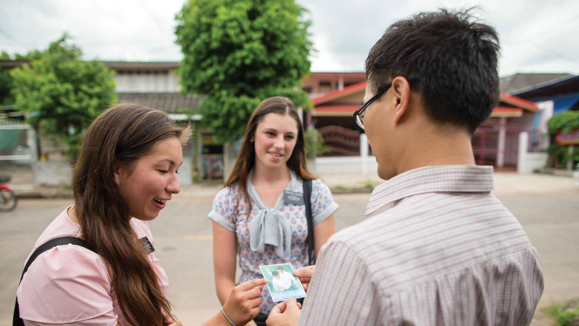 A photo of a pair of sister missionaries in Thailand speak to a man on the street and handing him a passalong card